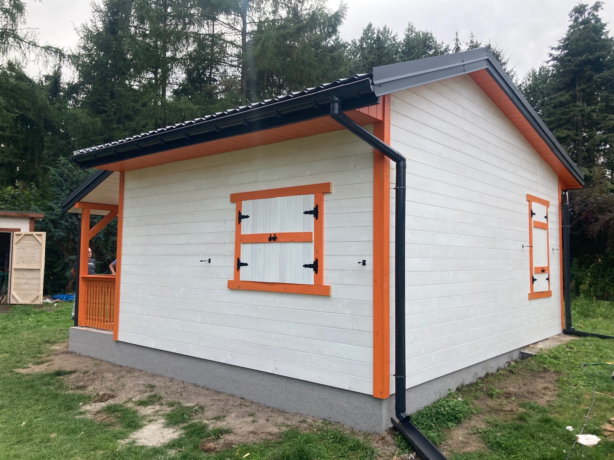 Nowoczesny, parterowy budynek z białymi deskami i pomarańczowymi obramowaniami okien, z czarną blachodachówką i rynnami, otoczony zielenią.