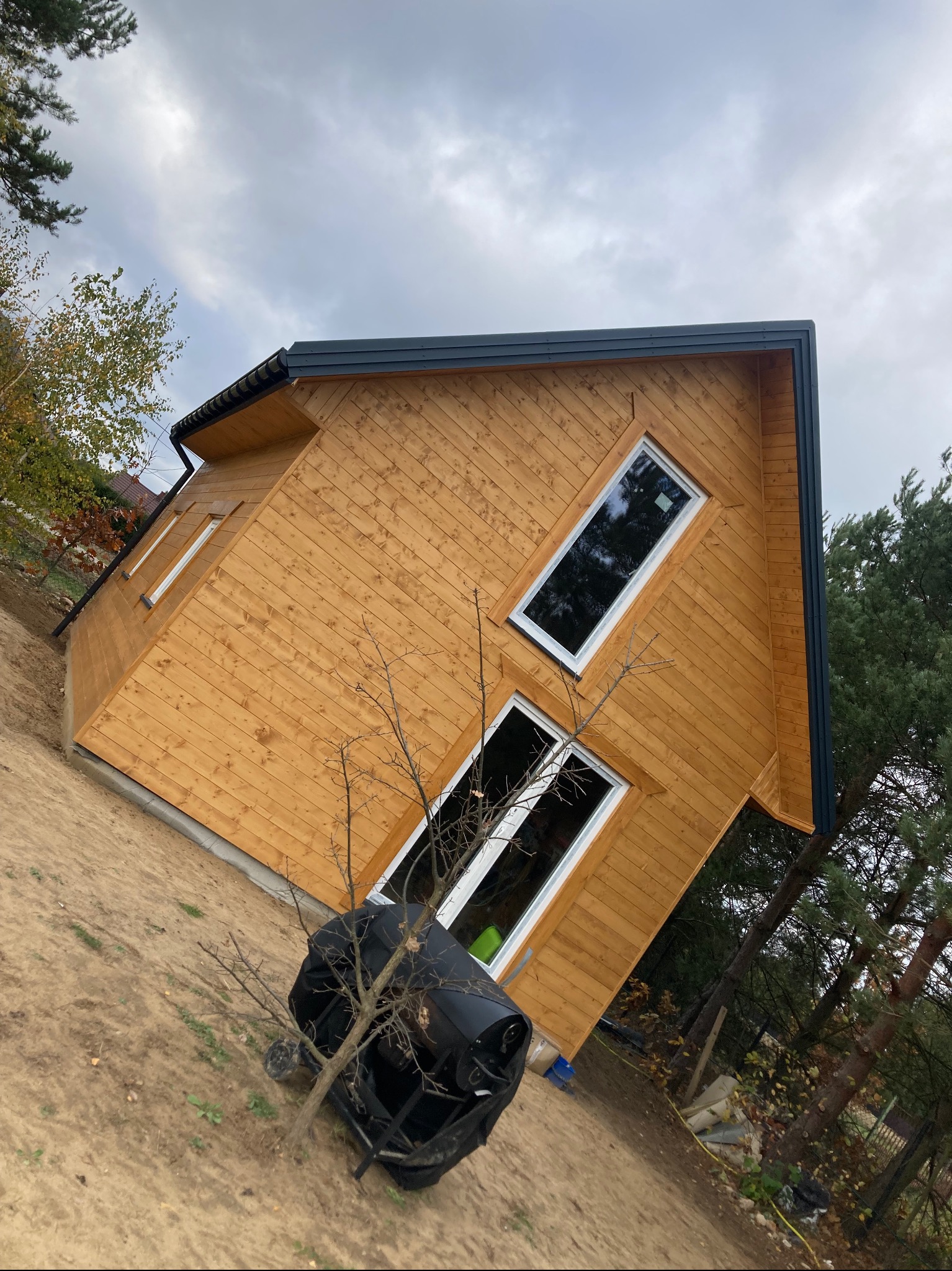 Surowy stan zewnętrzny małego, drewnianego domu z oknami, na działce z piaskiem i młodymi drzewami, pod pochmurnym niebem.