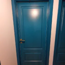 Turkusowe drzwi wewnętrzne z dwoma panelami, złotą klamką i ościeżnicą, widoczne na tle jasnej ściany i drewnianej podłogi, z fragmentem drugich drzwi po prawej stronie.