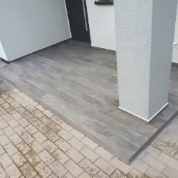 Zadaszone wejście do domu z szarymi panelami imitującymi drewno, wykończone białą listwą przy ścianie i kolumnie, oraz kostką brukową przed wejściem.