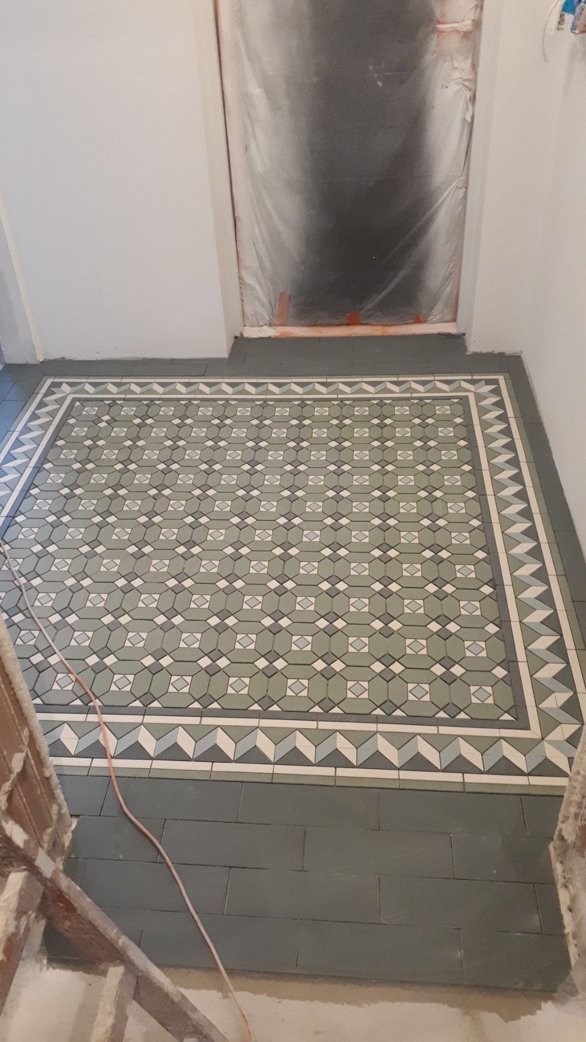 Ułożona podłoga z dekoracyjnych, geometrycznych płytek w odcieniach zieleni i szarości, z fragmentem zabezpieczonego folią wejścia i widocznym kablem na podłodze.