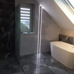 Nowoczesna łazienka na poddaszu z kabiną prysznicową typu walk-in, wolnostojącą białą wanną, szarą marmurową glazurą i oświetleniem LED w pionowych szczelinach.