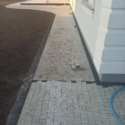 Jasnoszara kostka brukowa ułożona częściowo na ścieżce przy budynku, z widocznymi zapasowymi kostkami i fragmentem węża ogrodowego.