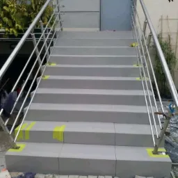 Zewnętrzne schody wyłożone szarymi płytami, z metalową balustradą, zabezpieczone żółtą taśmą malarską na krawędziach, prowadzące do szarego budynku.