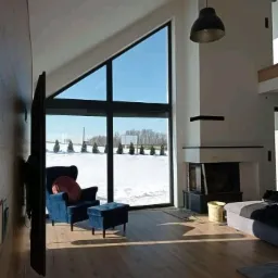 Wnętrze salonu z wysokim sufitem, dużym trójkątnym oknem z widokiem na zimowy krajobraz, nowoczesnym kominkiem i niebieskim fotelem z pufą.