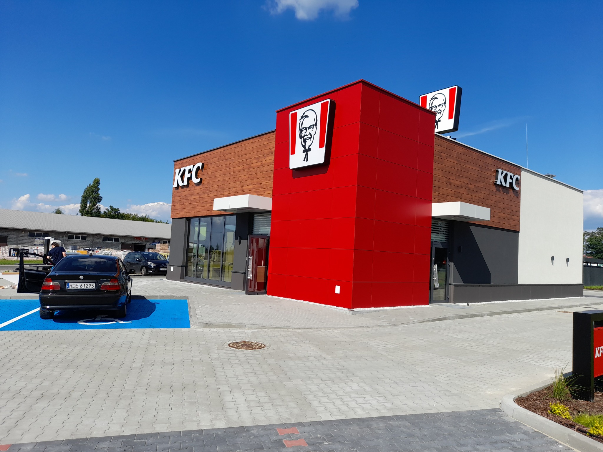Nowoczesny budynek restauracji KFC z elementami elewacji w kolorze czerwonym, brązowym i białym, logo na fasadzie i dachu, zaparkowany samochód na niebieskim miejscu parkingowym, kostka brukowa...