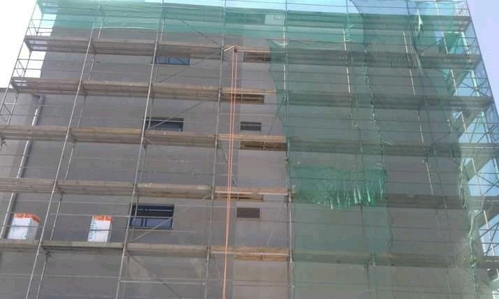 Budynek w trakcie prac elewacyjnych, osłonięty siatką ochronną i otoczony rusztowaniem, widoczne okna i materiały budowlane na rusztowaniu.