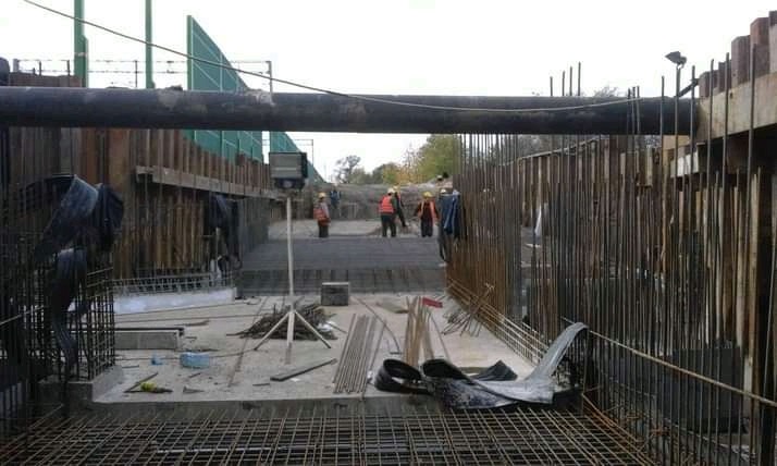 Budowa fundamentów: zbrojenie, szalunki, robotnicy w odblaskowych kamizelkach w tle. Widoczne pręty zbrojeniowe, drewniane elementy szalunków i duża rura nad konstrukcją.