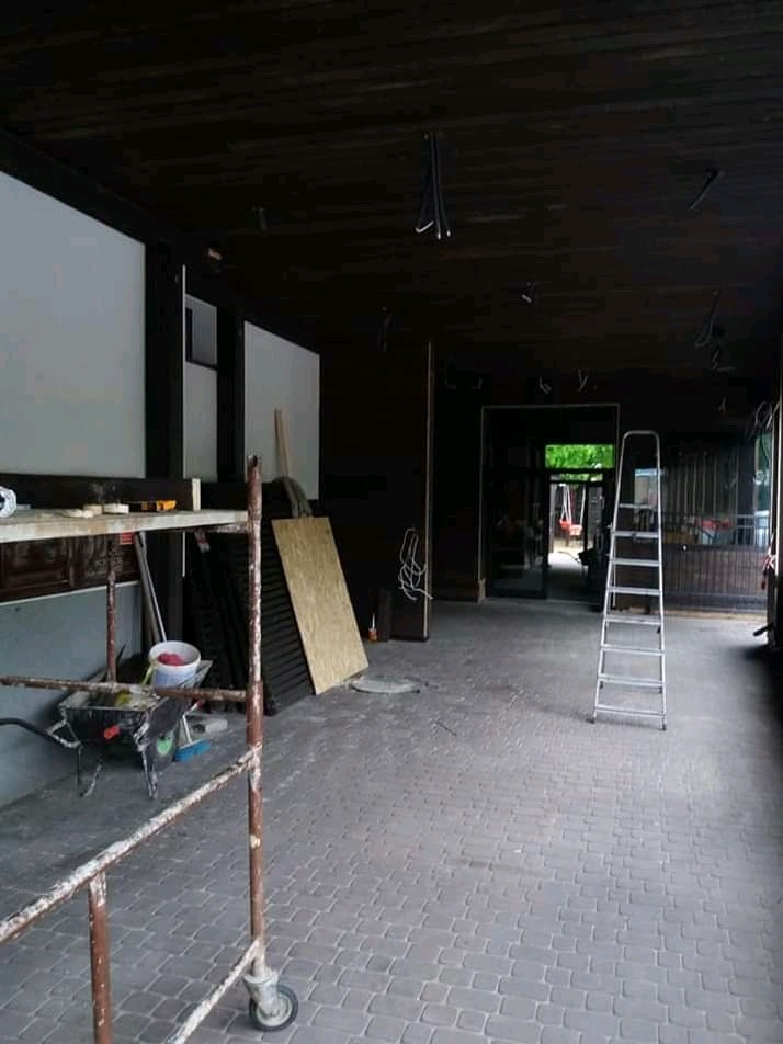 Wnętrze w trakcie remontu: widoczne rusztowanie, drabina, płyty, wiadro z farbą i taczka na tle ciemnego sufitu z wiszącymi kablami.