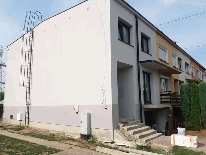 Świeżo pomalowany fragment elewacji szeregowca w odcieniach bieli i szarości, z widoczną drabiną przeciwpożarową na ścianie szczytowej i pozostałościami materiałów budowlanych przed budynkiem.