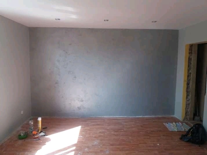 Świeżo pomalowane szare ściany z efektem dekoracyjnym w pustym pokoju z drewnianą podłogą, widoczne narzędzia malarskie i torba robocza.