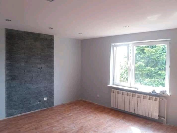 Pokój po remoncie: szare ściany, drewniana podłoga, okno z widokiem na drzewa i dekoracyjna ściana z szarej cegły.