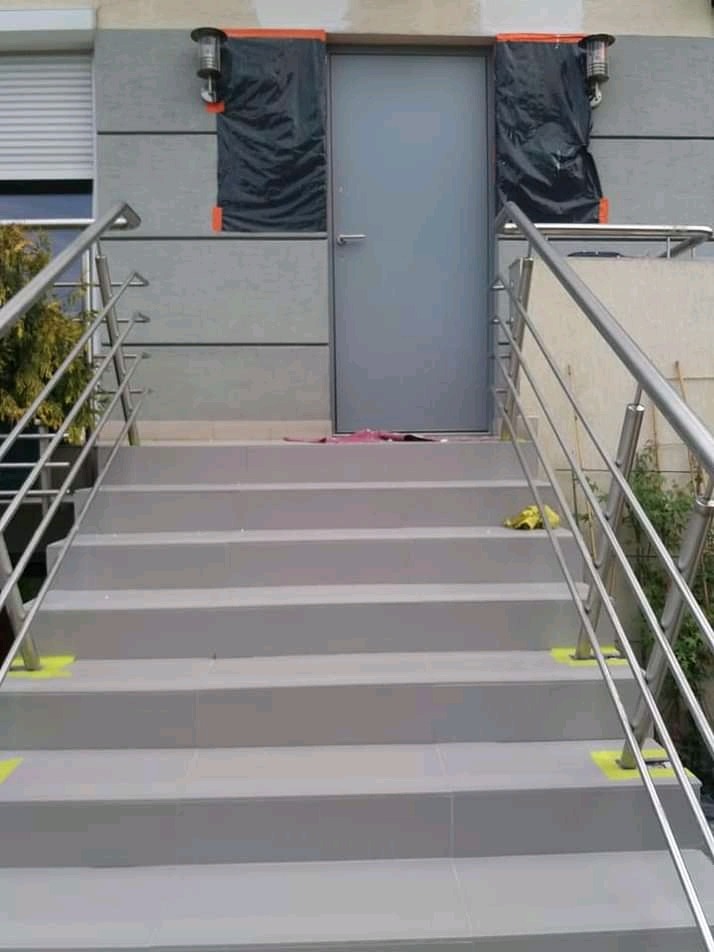 Zewnętrzne schody wyłożone szarymi płytkami prowadzące do drzwi wejściowych, zabezpieczone lampy i fragment elewacji. Balustrada ze stali nierdzewnej po obu stronach.