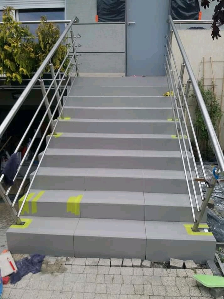 Zewnętrzne schody wyłożone szarymi płytami, z metalową balustradą, zabezpieczone żółtą taśmą malarską na krawędziach, prowadzące do szarego budynku.
