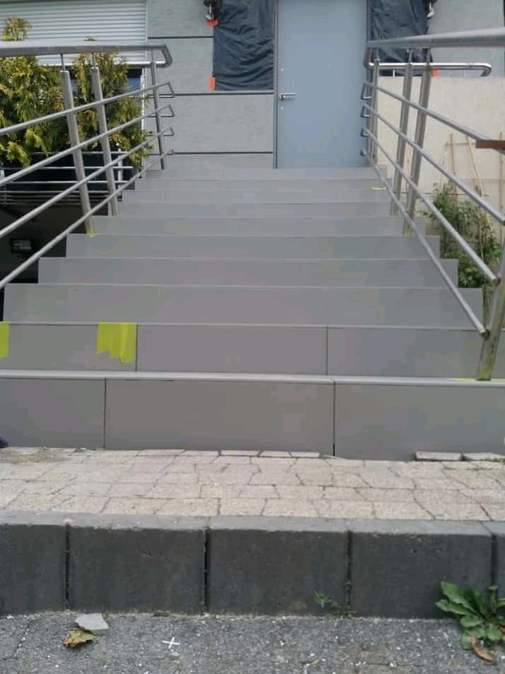 Schody zewnętrzne prowadzące do drzwi wejściowych, świeżo pomalowane na szaro, z metalowymi barierkami i fragmentem taśmy malarskiej zabezpieczającej krawędź.