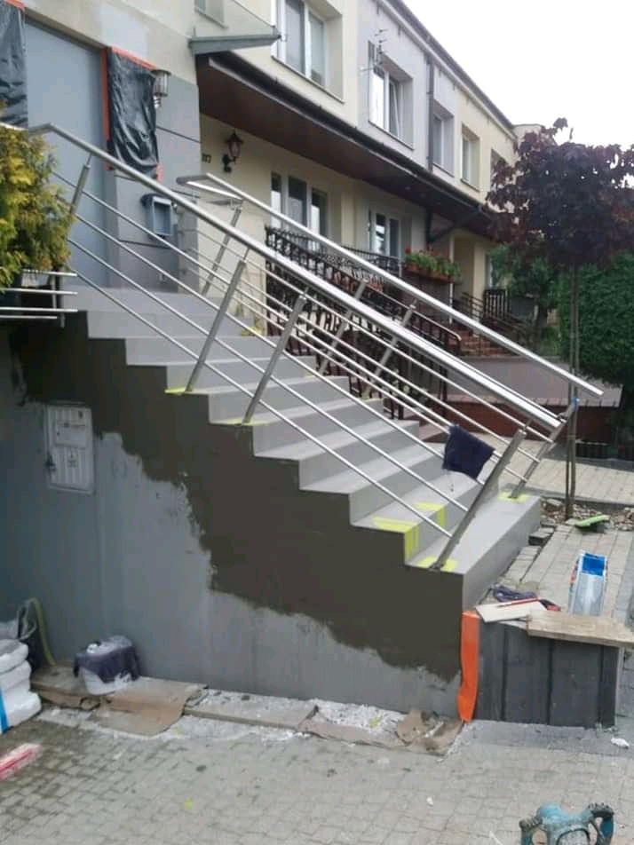 Zewnętrzne schody z metalową balustradą po remoncie, z widocznymi śladami prac na ścianie i stopniach, otoczone materiałami budowlanymi.