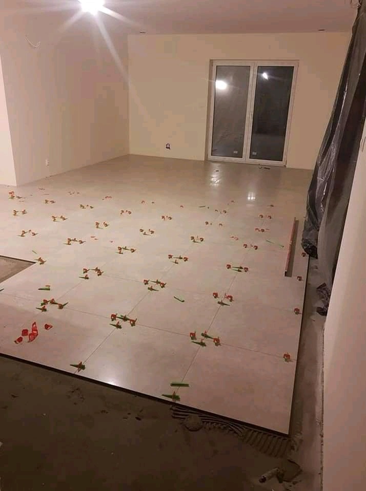 Układanie jasnych płytek podłogowych w dużym pokoju, widoczne krzyżyki dystansowe i poziomica, okno balkonowe w tle.