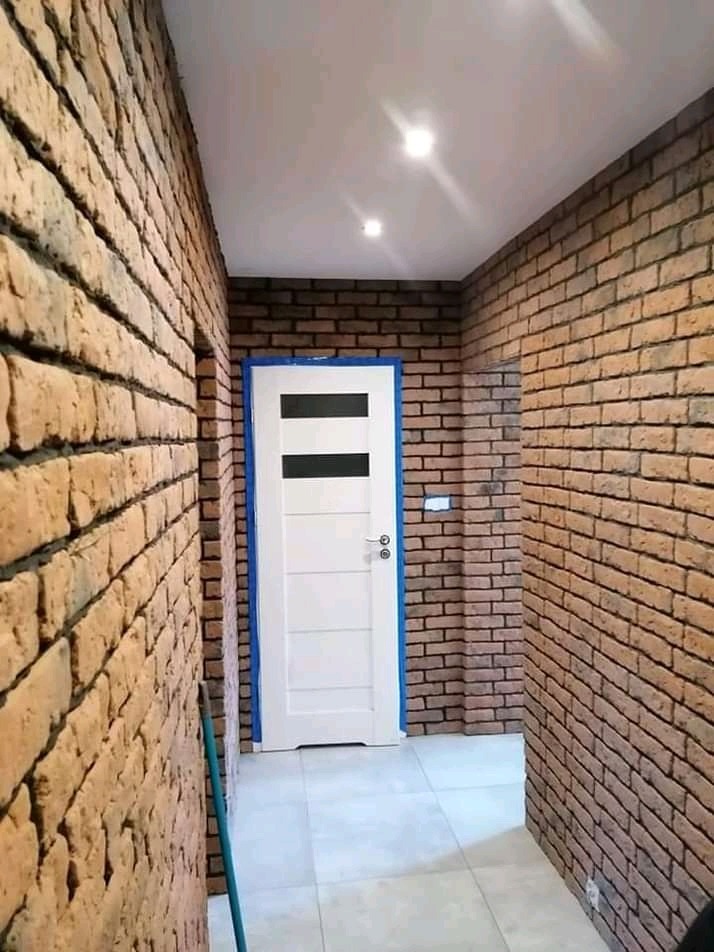 Korytarz wykończony cegłą klinkierową w odcieniach brązu, z białą, nowoczesną stolarką drzwiową zabezpieczoną niebieską taśmą malarską i jasną podłogą z płytek.