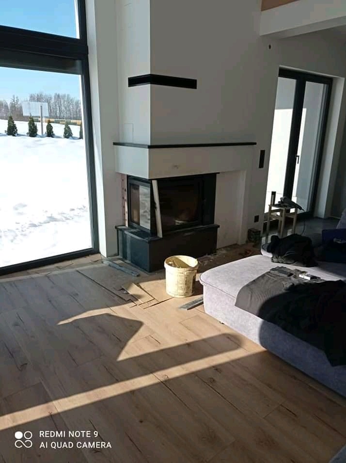 Wnętrze domu w trakcie wykańczania: obudowa kominka z czarnymi akcentami, fragment podłogi z paneli, widoczne okno z widokiem na zaśnieżony ogród, wiadro i elementy wykończeniowe na podłodze...