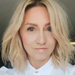 Portret uśmiechniętej kobiety z blond, falowanymi włosami i jasnymi oczami, ubrana w białą koszulę z ozdobnym guzikiem.