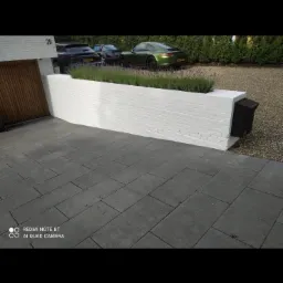 Biały, pomalowany murek z cegły z rosnącą na nim lawendą, skrzynka pocztowa, szara kostka brukowa na podjeździe, w tle zaparkowane samochody.