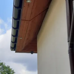 Kamera monitoringu zamontowana pod drewnianym okapem budynku z widocznymi rynnami i oświetleniem punktowym.