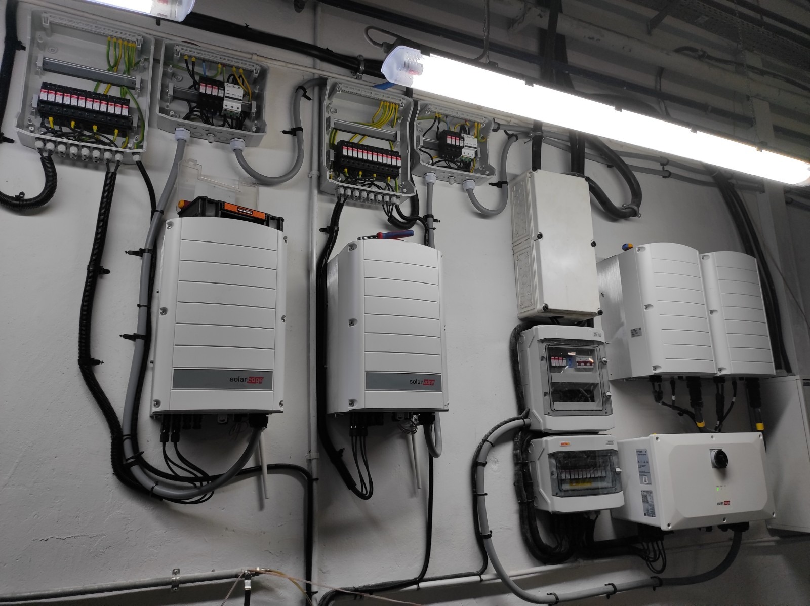 Instalacja fotowoltaiczna z widocznymi inwerterami SolarEdge i rozdzielnicami elektrycznymi na białej ścianie, oświetlona lampą jarzeniową.