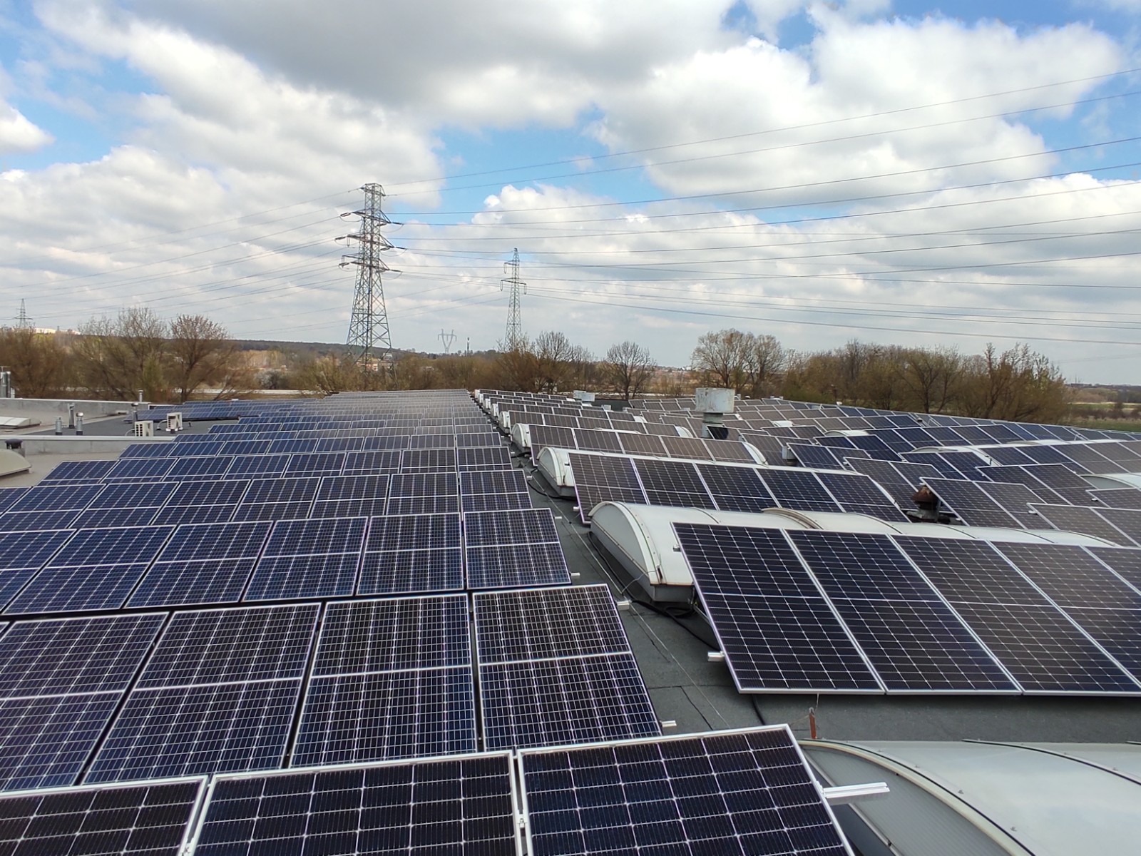 Rozległa instalacja paneli słonecznych na dachu przemysłowym, z widocznymi liniami wysokiego napięcia i błękitnym niebem w tle.