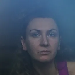 Portret kobiety z kręconymi włosami i w różowej koszulce na tle ciemnego otoczenia, oświetlona od przodu.