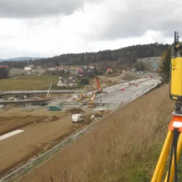 Żółto-szary tachimetr na statywie na tle budowy drogi ekspresowej w pagórkowatym terenie, widoczne maszyny budowlane i fragmenty konstrukcji mostu.