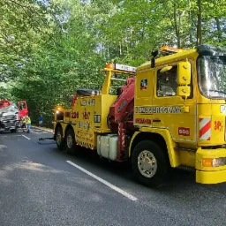 Żółta laweta z napisem 'Pomoc Drogowa' na leśnej drodze w Jełowej, holująca uszkodzoną ciężarówkę po wypadku, z pracownikami zabezpieczającymi pojazdy.