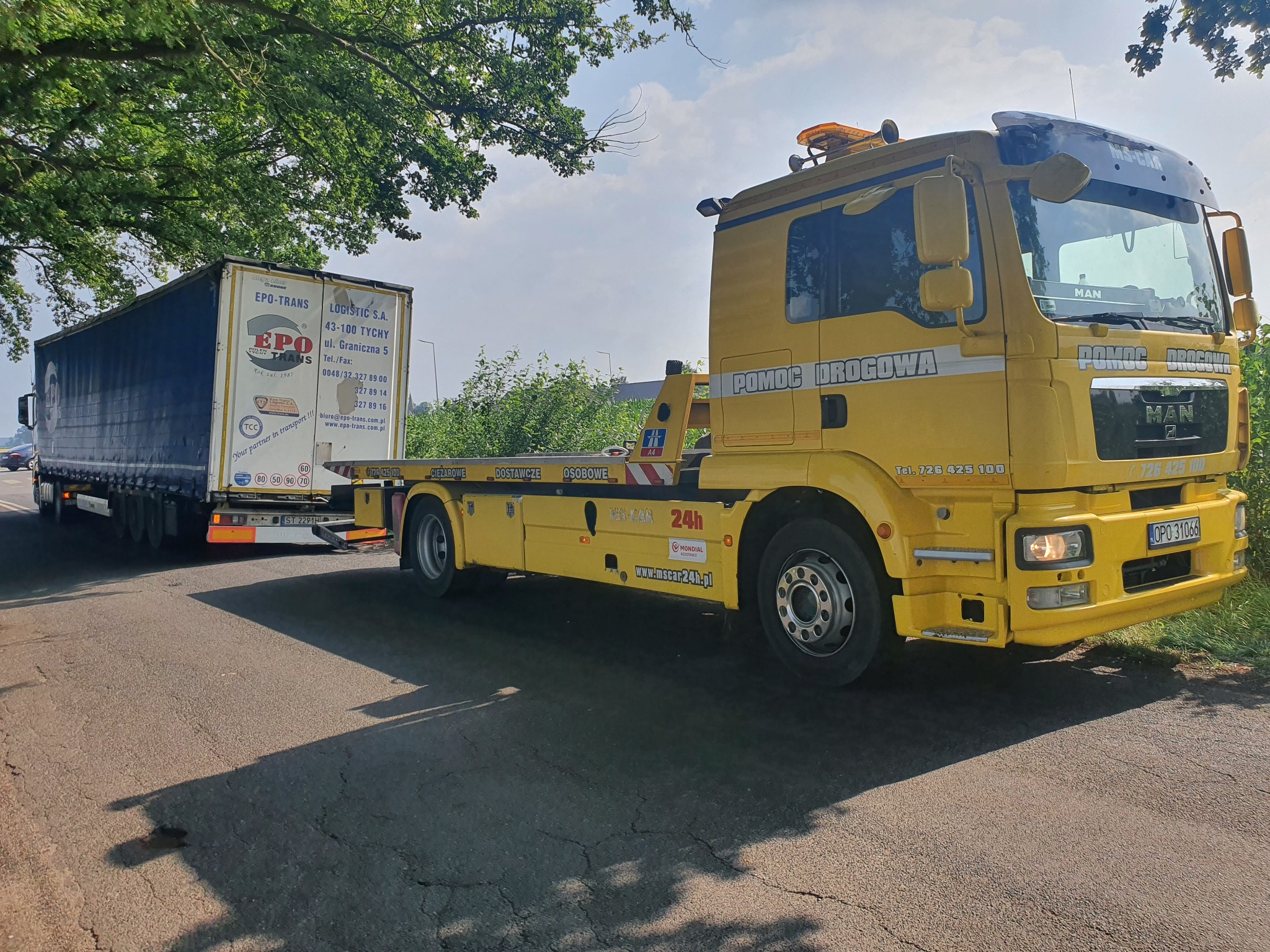 Żółta laweta z napisem 'Pomoc Drogowa' firmy z Jełowej holuje naczepę z logo EPO-TRANS na tle zieleni i błękitnego nieba.