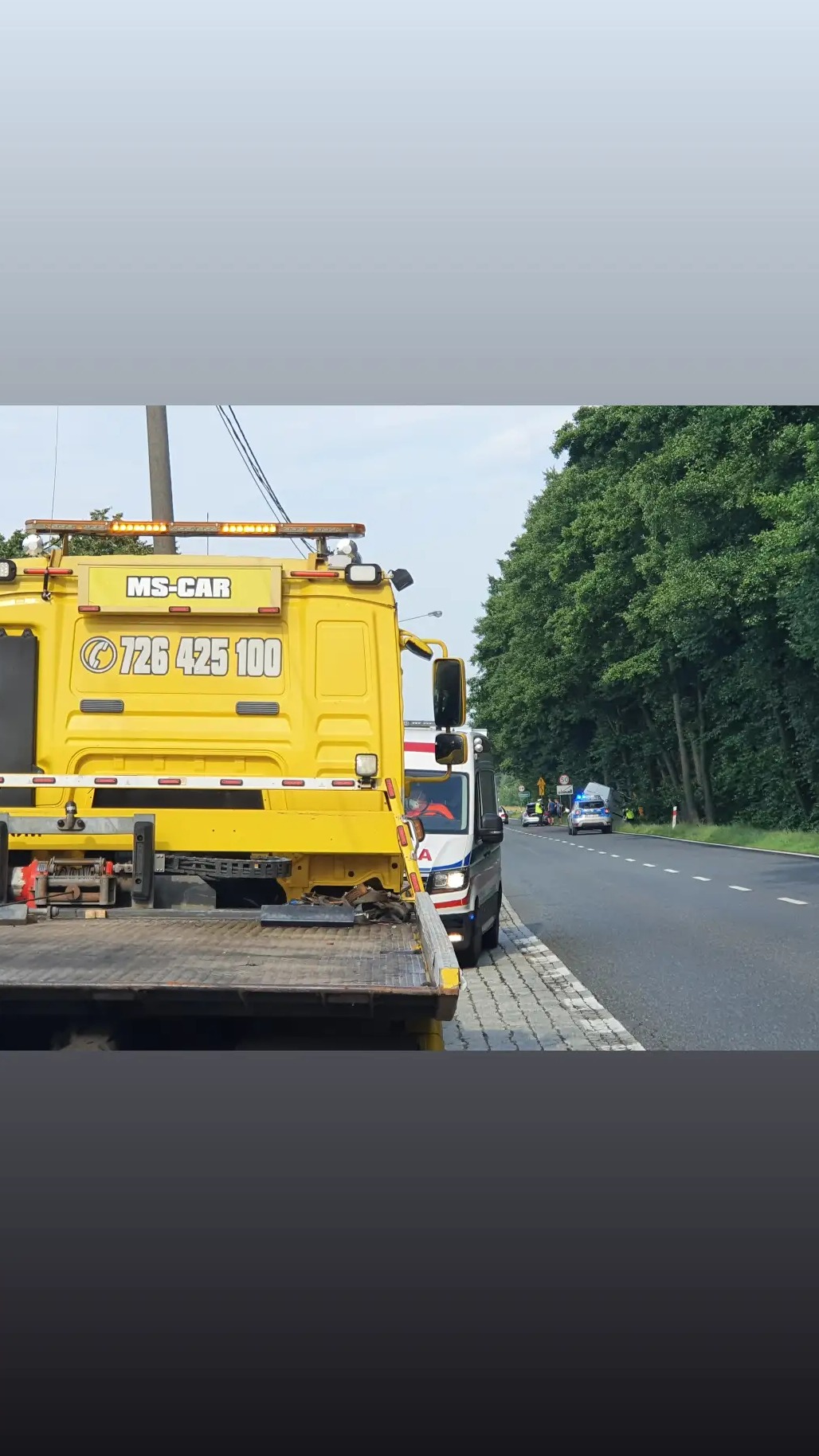 Żółta laweta MS-CAR z numerem telefonu 726 425 100 na drodze, obok ambulansu, w tle policyjny radiowóz i grupa osób.