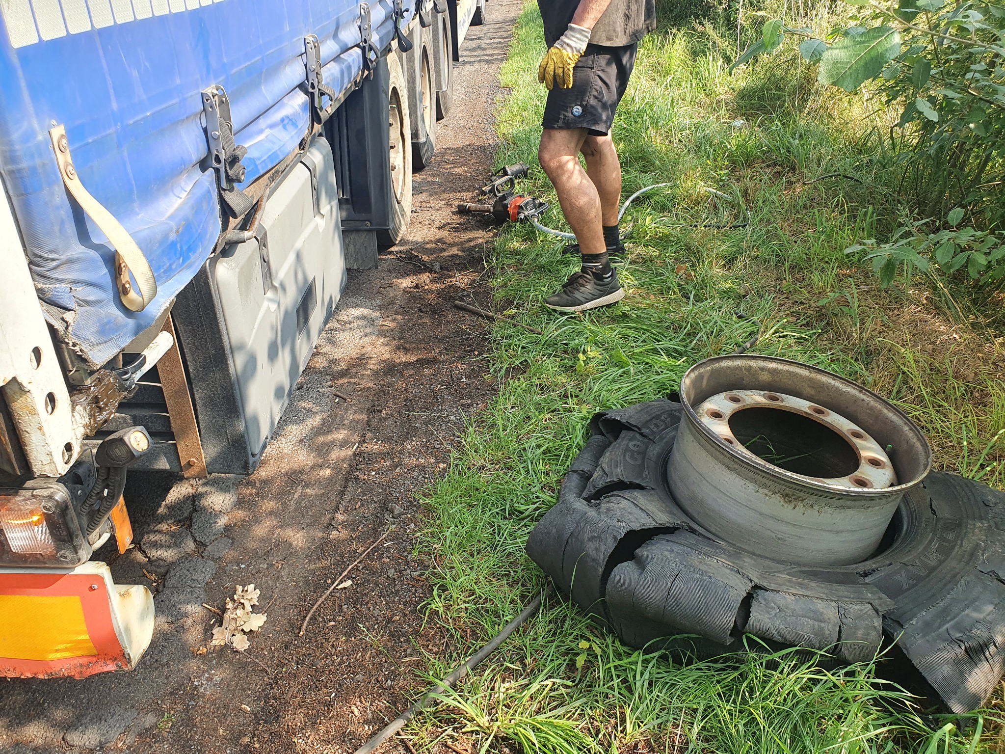 Uszkodzone koło ciężarówki z felgą leżące na trawie, obok stoi ciężarówka i narzędzia do wymiany kół, widoczne nogi mechanika w roboczych rękawicach.