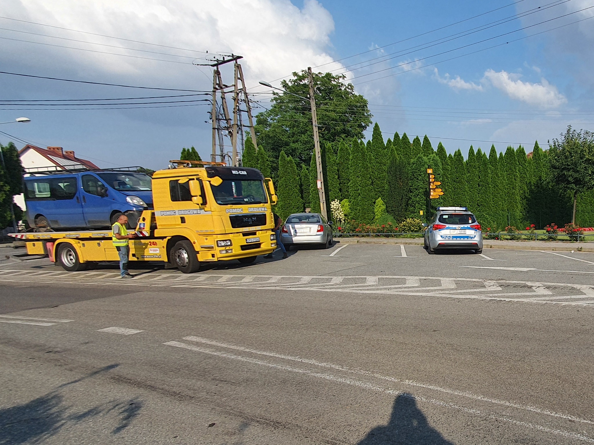 Żółta laweta MAN z niebieskim busem na platformie, pracownik w kamizelce odblaskowej stoi obok, na parkingu srebrny samochód osobowy i radiowóz policyjny, w tle zielone tuje i słup energetyczny.