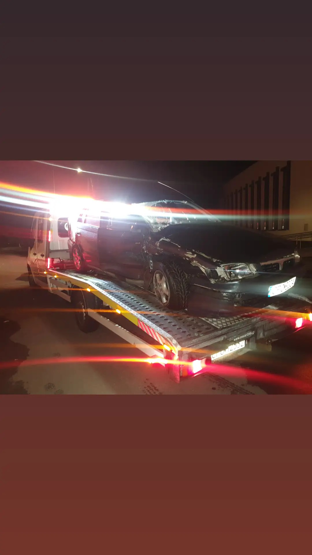 Uszkodzony, czarny samochód osobowy z rozbitym przodem i szybą, załadowany na lawetę oświetloną czerwonymi światłami w nocy.