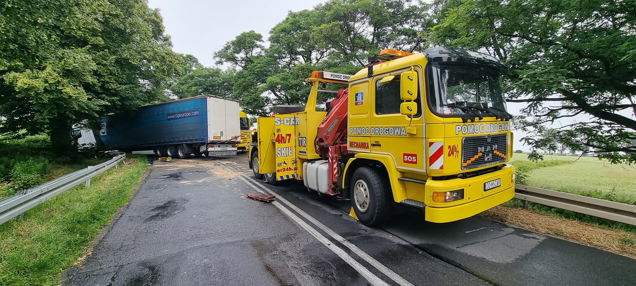 Żółta laweta pomocy drogowej MAN z wysięgnikiem, na mokrej drodze, zabezpieczona klinem pod kołem, holuje uszkodzoną ciężarówkę, która uderzyła w drzewo przy drodze. Na asfalcie leżą fragmenty...