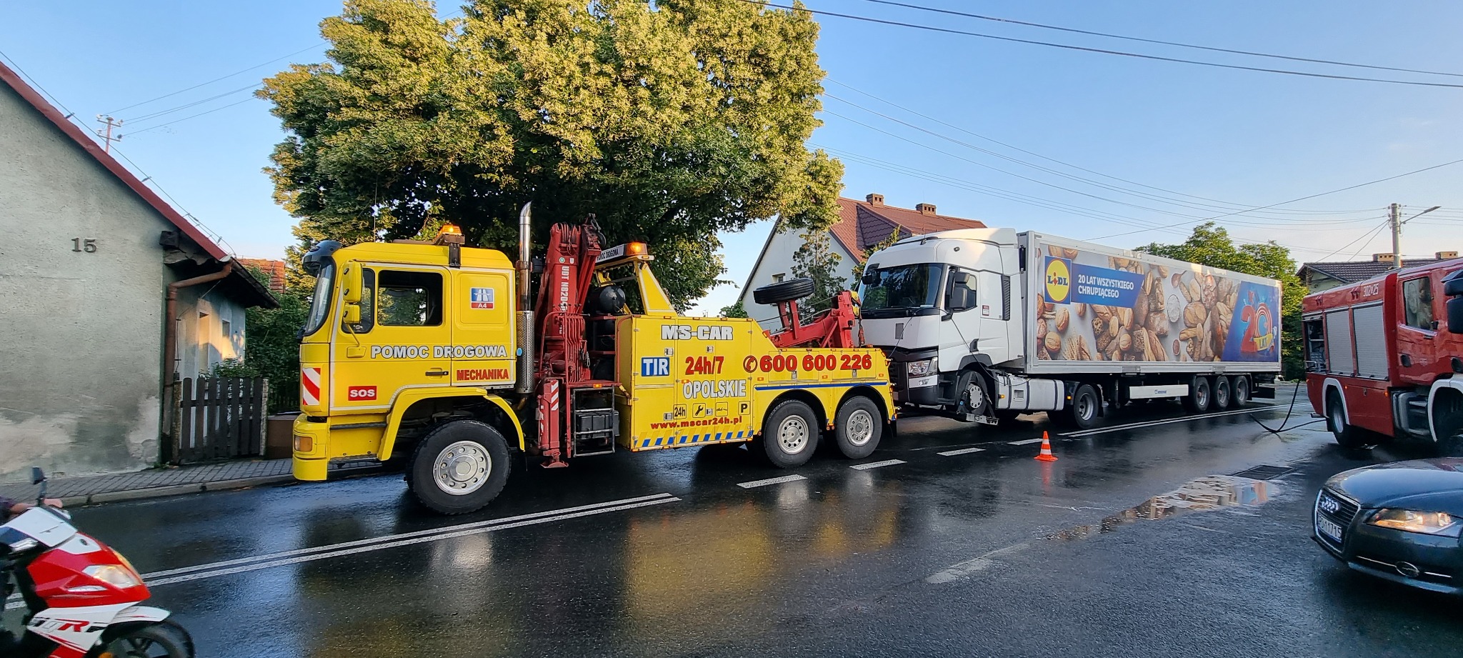 Żółta laweta z napisem 'Pomoc Drogowa Mechanika' holuje białą ciężarówkę z reklamą Lidl na mokrej ulicy, w tle budynki mieszkalne i wóz strażacki.