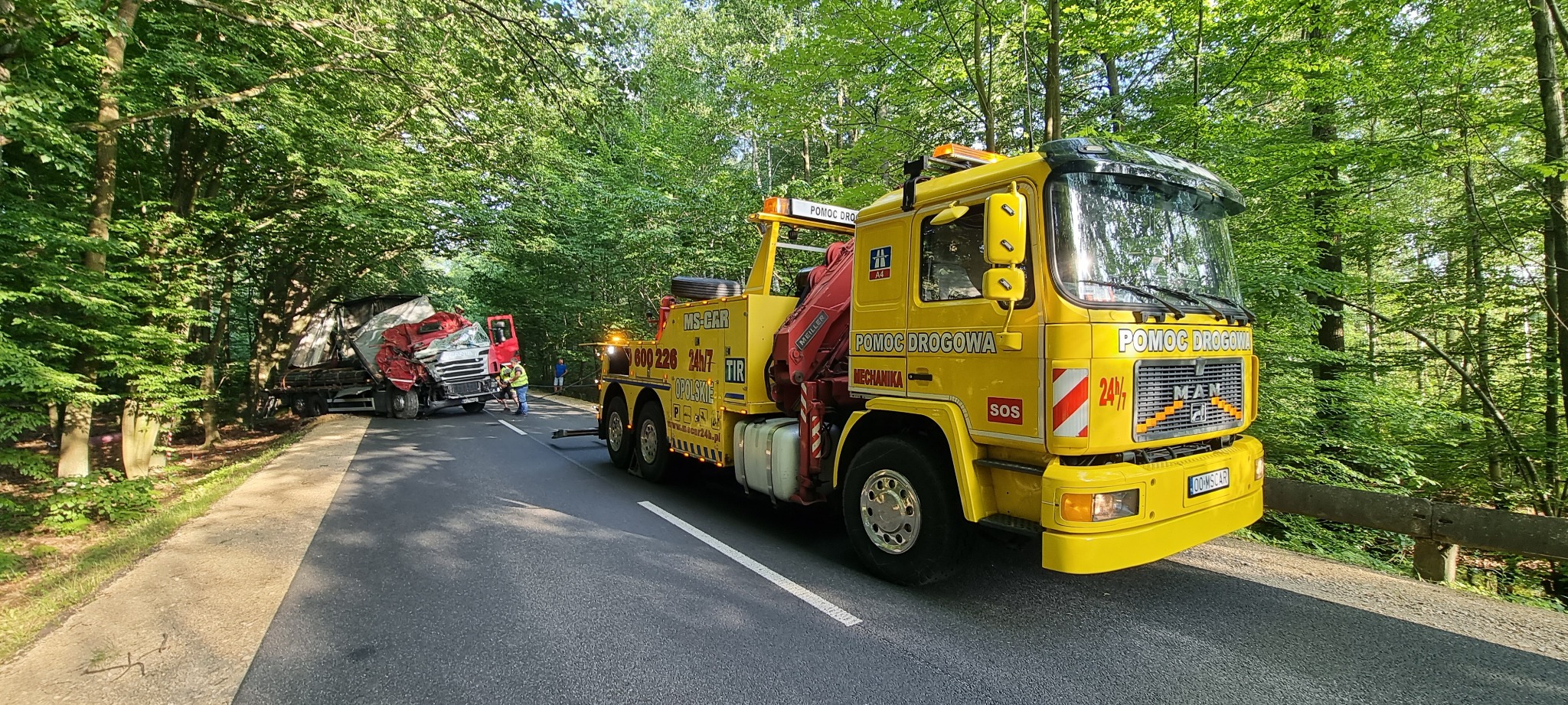 Żółta laweta z napisem 'Pomoc Drogowa' na leśnej drodze w Jełowej, holująca uszkodzoną ciężarówkę po wypadku, z pracownikami zabezpieczającymi pojazdy.