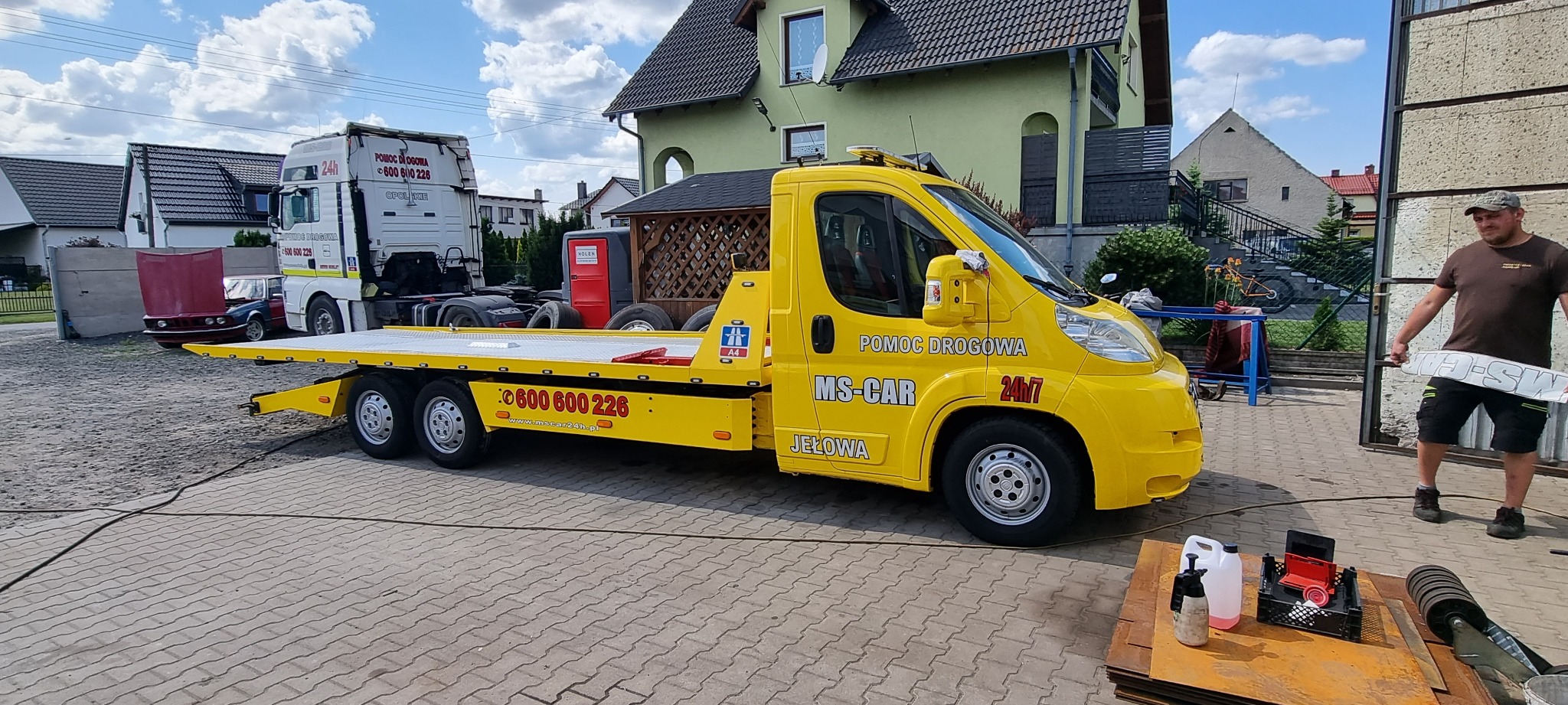 Żółta laweta z napisem 'POMOC DROGOWA MS-CAR JEŁOWA 24h/7' na tle domu i zaparkowanego tira. Mężczyzna w czapce trzymający element z napisem 'MS-CAR' obok.