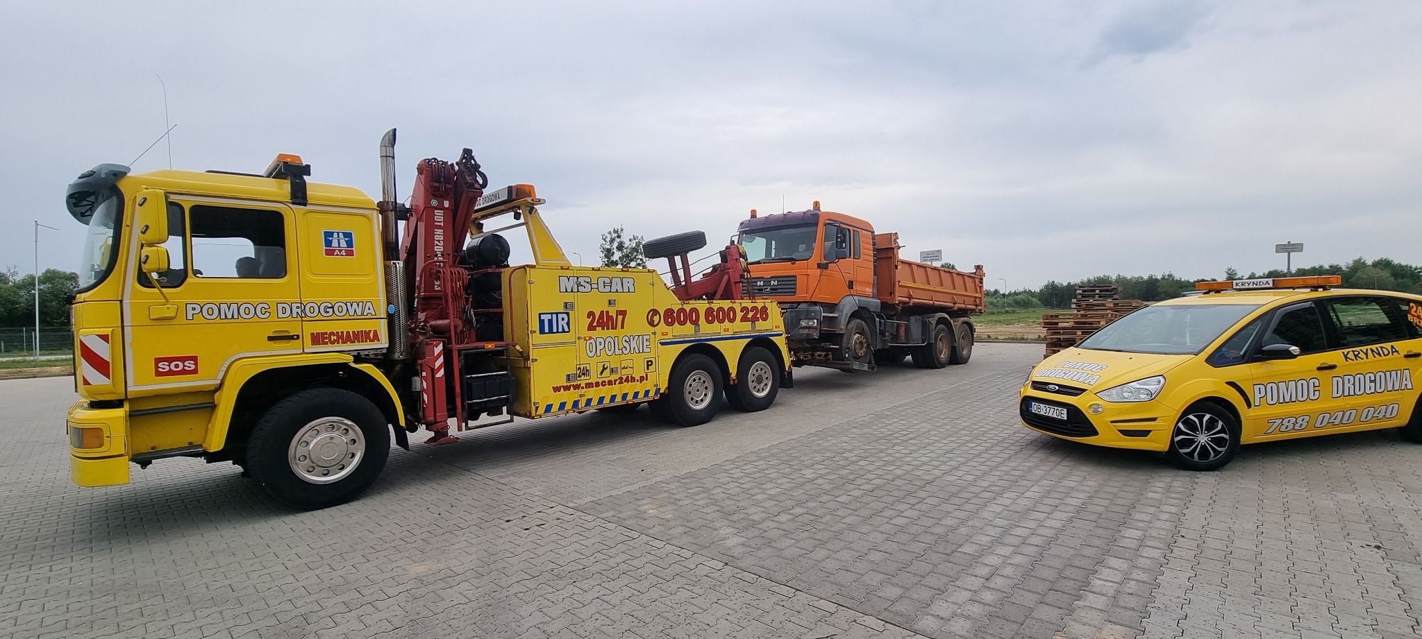 Żółta laweta z dźwigiem holuje pomarańczową ciężarówkę wywrotkę, obok stoi żółty samochód osobowy z napisem Pomoc Drogowa na parkingu z kostki brukowej.
