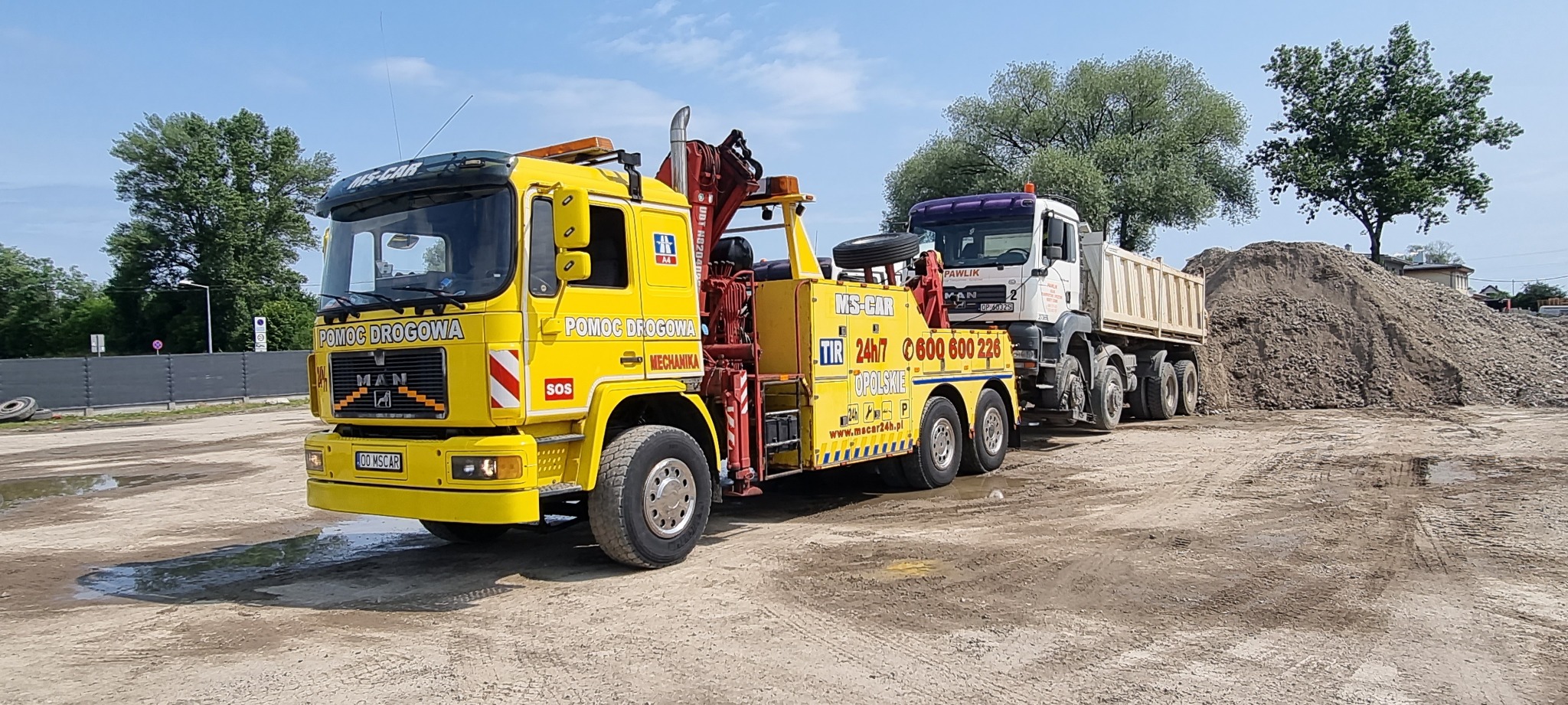 Żółta laweta MAN z napisem 'Pomoc Drogowa' holuje ciężarówkę z przyczepą na piaszczystym placu, w tle hałda piasku i drzewa.