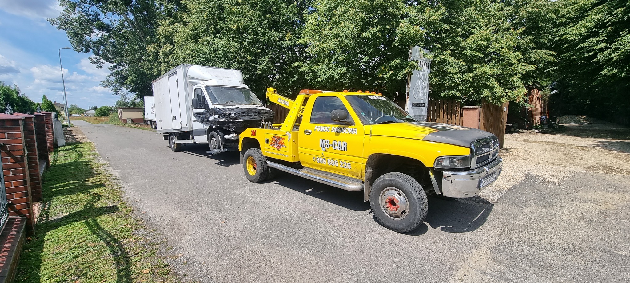 Żółta laweta holuje uszkodzony biały samochód dostawczy na asfaltowej drodze w słoneczny dzień.