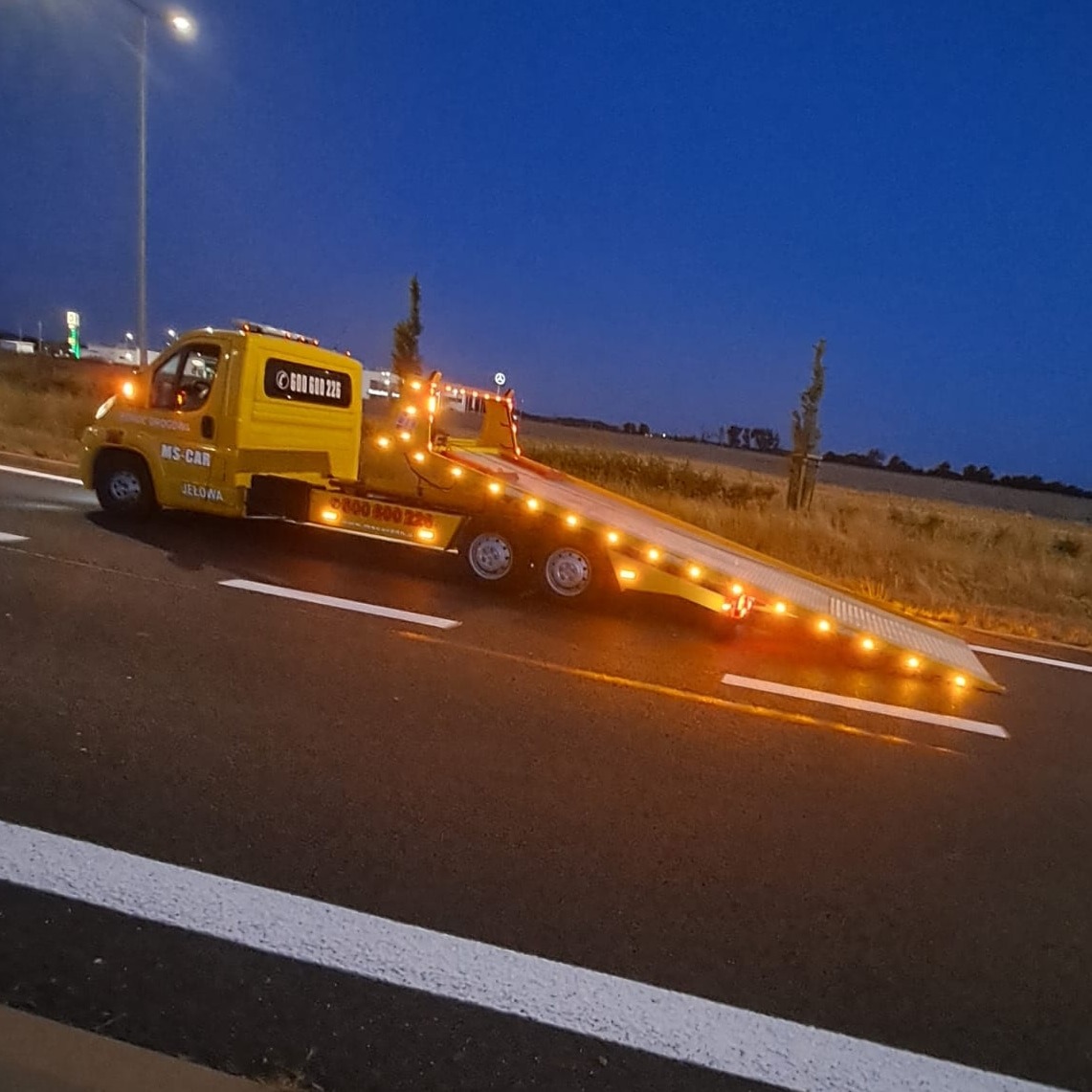 Żółta laweta MS-CAR z włączonymi światłami ostrzegawczymi, stojąca na poboczu drogi o zmierzchu, widoczne numery kontaktowe na kabinie.