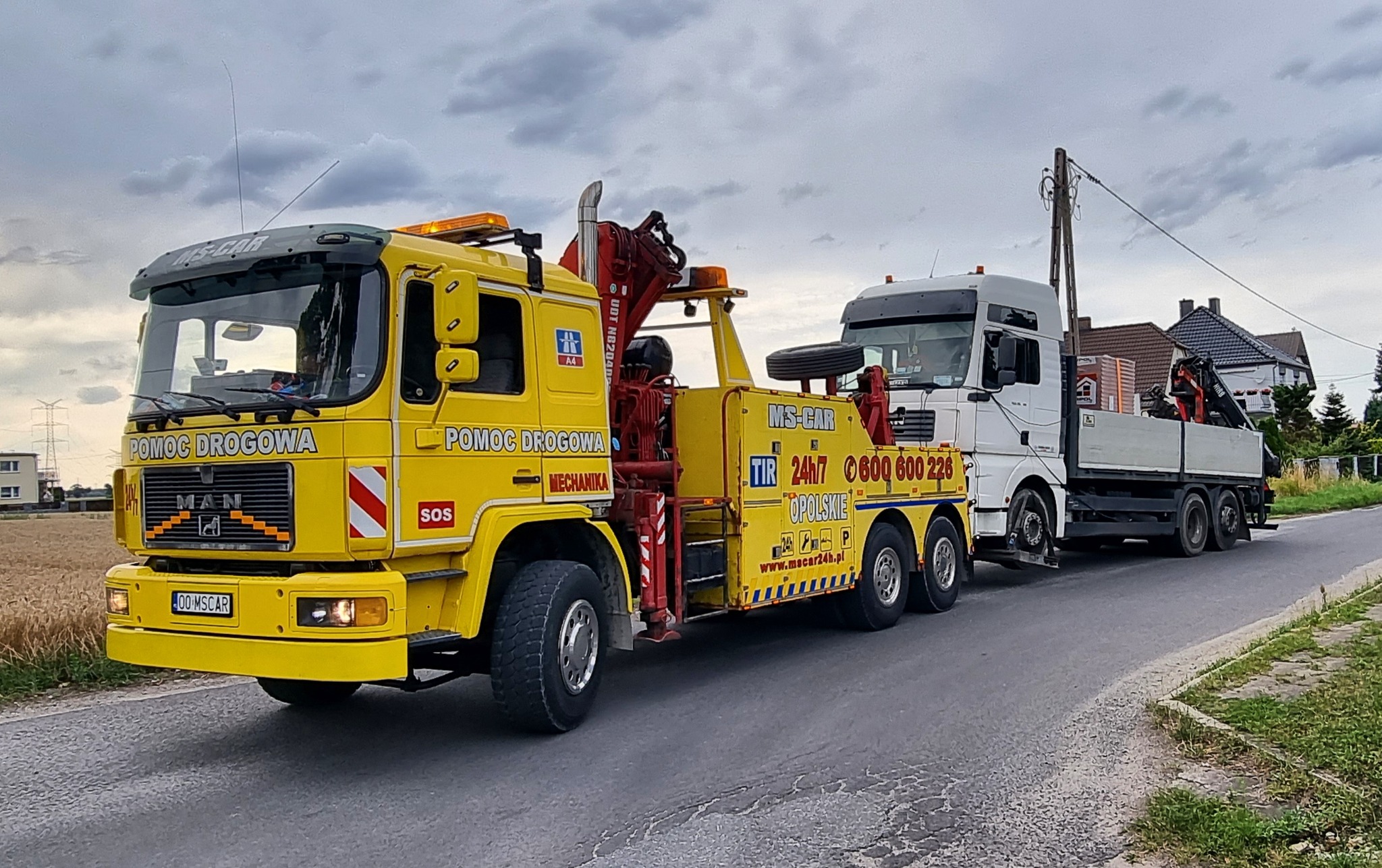 Żółta laweta MAN z napisem 'Pomoc Drogowa' holuje białą ciężarówkę z ładunkiem na drodze asfaltowej, pochmurne niebo w tle.