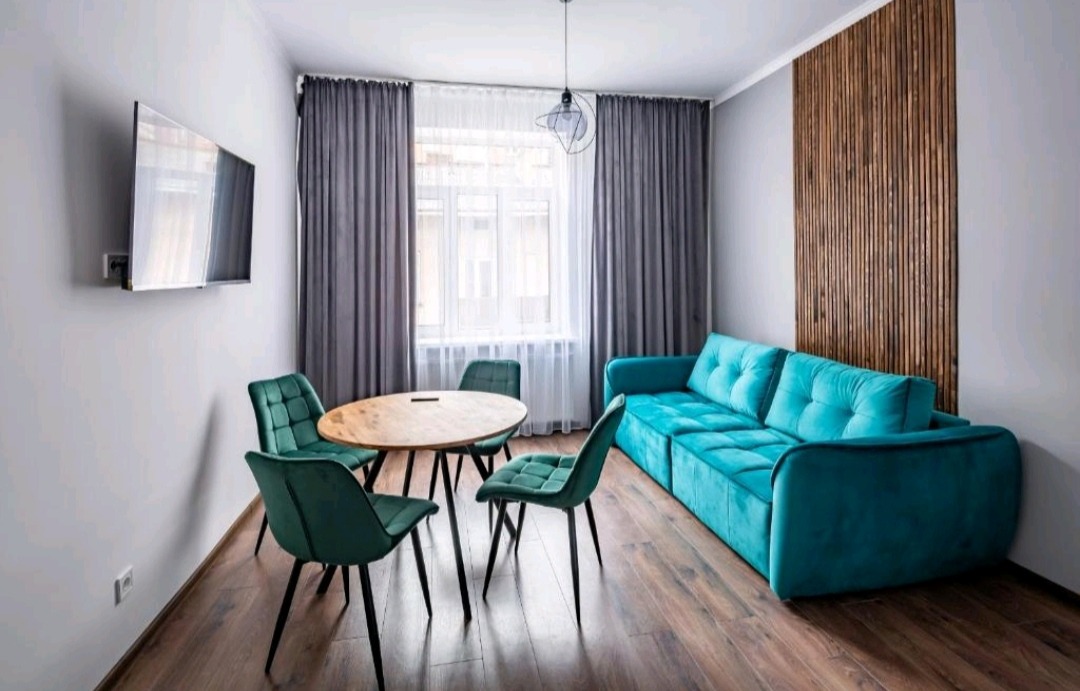 Jasne wnętrze pokoju dziennego z turkusową sofą, okrągłym drewnianym stołem otoczonym przez cztery zielone krzesła na tle drewnianej ściany i szarych zasłon.