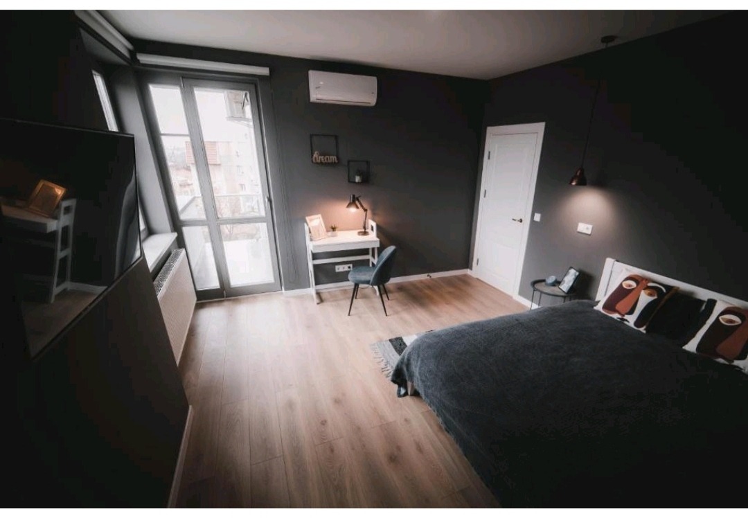 Nowoczesna sypialnia z szarymi ścianami, drewnianą podłogą i minimalistycznym biurkiem przy oknie, oświetlona lampą sufitową i biurkową.