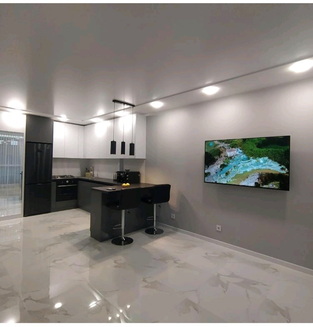 Nowoczesna, czarno-biała kuchnia z wyspą, trzema wiszącymi lampami i telewizorem na ścianie, podłoga wyłożona marmurowymi płytkami.