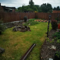 Ogród w trakcie transformacji: widoczne fundamenty pod taras, gruz po wyburzeniach, metalowa rynna na trawie, lampy ogrodowe wzdłuż ścieżki, oraz granica trawnika wyłożona korą.