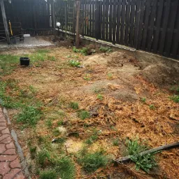 Przygotowany teren pod trawnik: usunięte stare rośliny, nawieziona ziemia, rozsypane igliwie, widoczne narzędzia ogrodnicze i fragment altanki w tle.